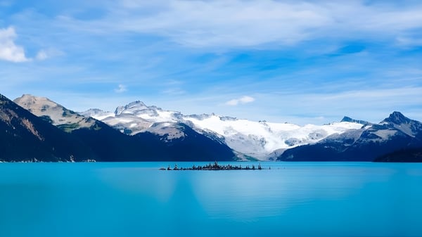 Ein klarer, türkisfarbener See vor schneebedeckten Bergen unter blauem Himmel, nahe der St. Benedict Catholic Secondary School.