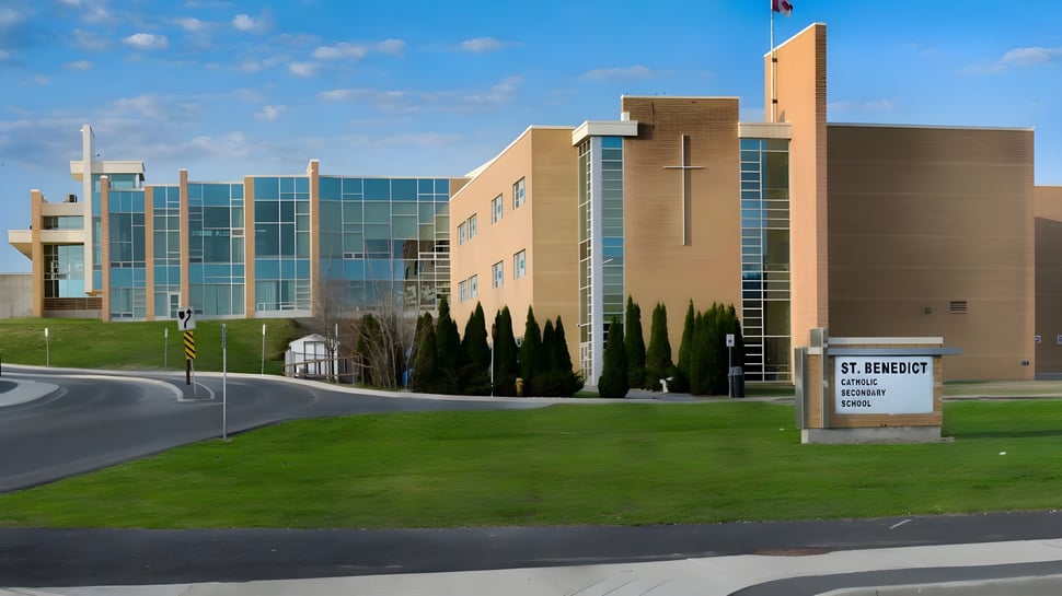 Das moderne mehrstöckige Schulgebäude der St. Benedict Catholic Secondary School mit Glasfassade und gepflegtem Vorgarten unter blauem Himmel.