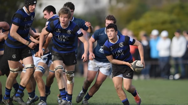 Schüler des St. Benildus College spielen Rugby auf einem grünen Spielfeld vor einer Sportanlage.