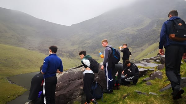 Schüler von St. Brendan's College stehen in Sportkleidung auf einem Felsen mit Blick auf eine neblige Berglandschaft.