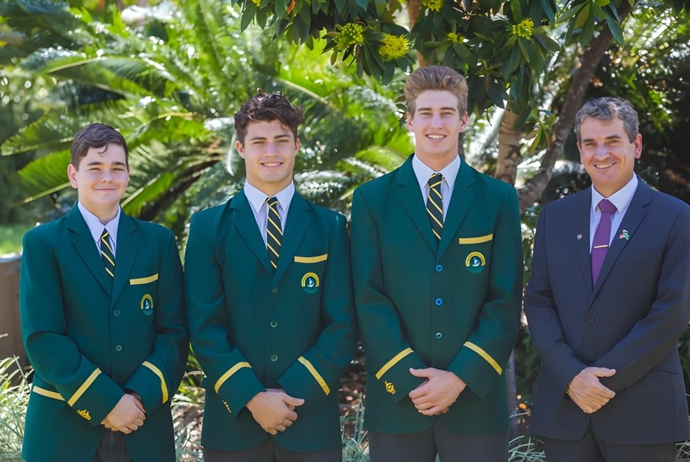 Vier Schüler des St. Brendan's College stehen in grünen und violetten Anzügen in einem tropischen Außenbereich mit Palmen.