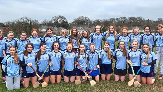 Eine Gruppe junger Athletinnen in blauen und weißen Uniformen steht auf dem Sportfeld des St. Brigid's College vor einem Waldgebiet.