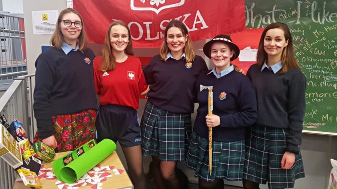 Eine Gruppe von Schülerinnen in Schuluniformen steht vor der Tafel mit verschiedenen Schriftzügen und der polnischen Flagge am St. Brigid's College.