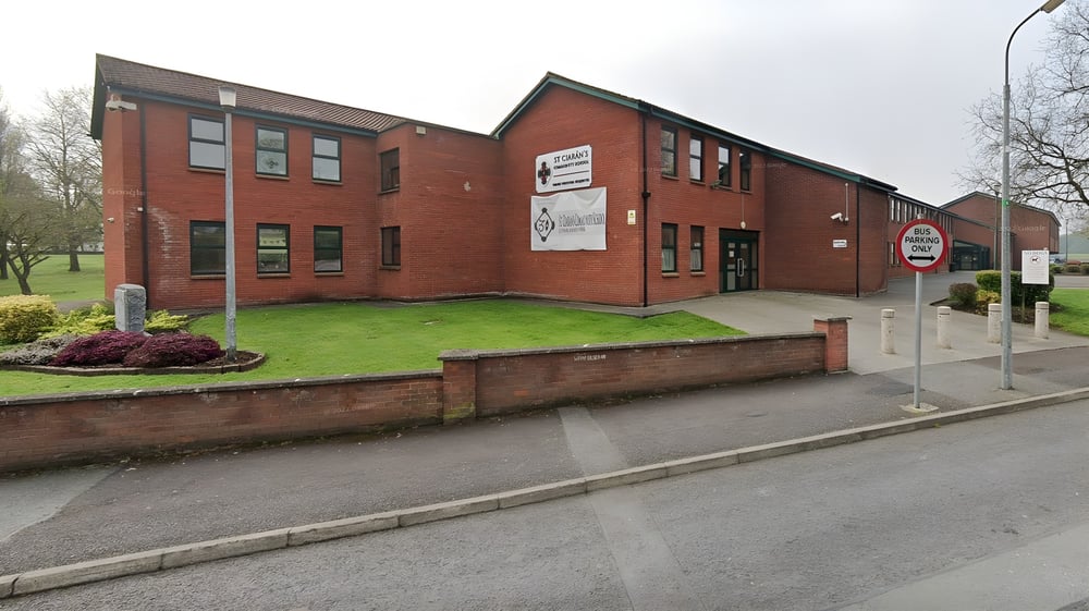 Das zweigeschossige Backsteingebäude der St. Ciarán’s Community School mit mehreren Fenstern steht auf einer Rasenfläche vor Bäumen und bewölktem Himmel.