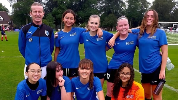 Eine Gruppe von Schülerinnen der St. Clare’s School in blauen Sportanzügen steht und sitzt auf einem Sportplatz mit Gras und Bäumen.