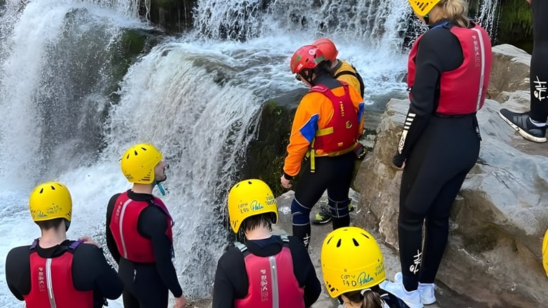 Eine Gruppe Schülerinnen und Schüler der St. Clare’s School steht in bunter Schutzkleidung vor einem rauschenden Wasserfall.