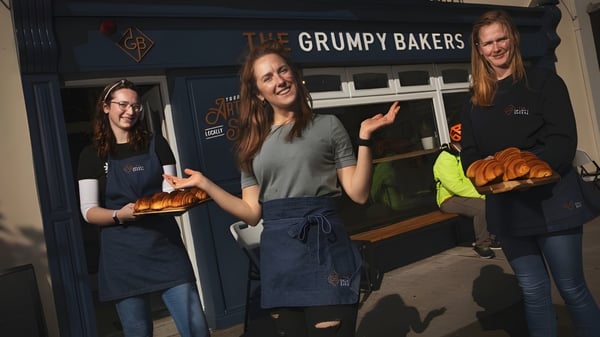 Drei Schülerinnen des St. Colman’s Community College stehen vor dem Laden The Grumpy Bakers und halten frisch gebackene Waren.