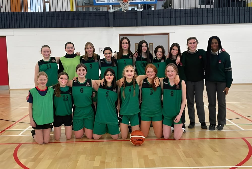 Eine Gruppe junger Basketballspielerinnen der St. Colman’s Community College posiert gemeinsam auf dem Basketballfeld.