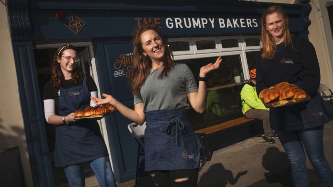 Drei Schülerinnen von St. Colman’s Community College stehen vor der Bäckerei The Grumpy Bakers und halten frisch gebackene Brote.