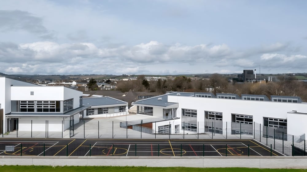 Das mehrstöckige Gebäude von St. Colman’s Community College mit Basketballplatz und hügeliger Stadtkulisse im Hintergrund.