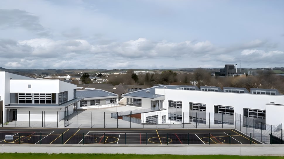 Das moderne Gebäude des St. Colman’s Community College steht vor einer hügeligen Stadtlandschaft mit großem Parkplatz im Vordergrund.