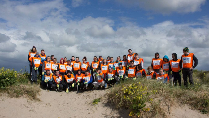Eine Gruppe von Schülern des St. Colman’s Community College steht mit orangen Sicherheitswesten auf einem Grasfeld vor bewölktem Himmel.