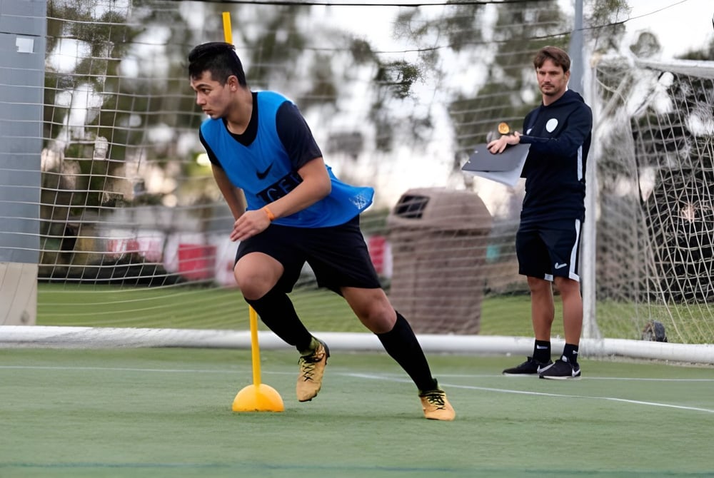 Ein Schüler des St. David's Catholic Sixth Form College dribbelt einen Fußball auf einem Rasenfeld, während eine weitere Person im Hintergrund zuschaut.