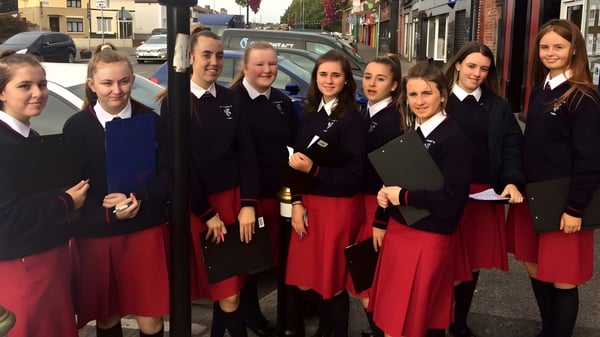 Schülerinnen von St. Dominic’s College stehen in Schuluniform auf einer Straßenszene mit Gebäuden und Fahrzeugen im Hintergrund.