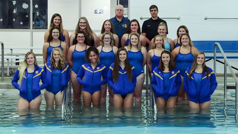 Eine Gruppe von jungen Schwimmerinnen in blauen Badeanzügen im Pool auf dem Gelände von St. Dominic's Girl’s Catholic College mit Trainer im Hintergrund.