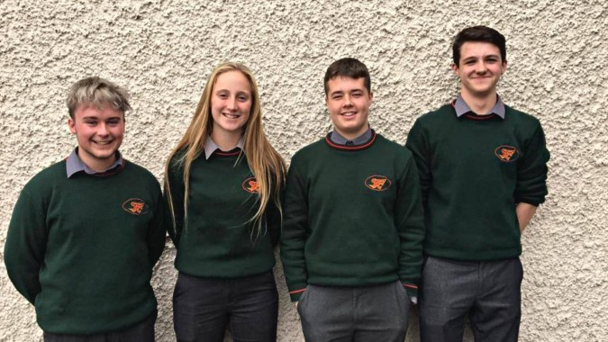 Vier Schüler in grünen Schuluniformen stehen vor einer strukturierten Wand auf dem Campus von St. Fergal’s College.