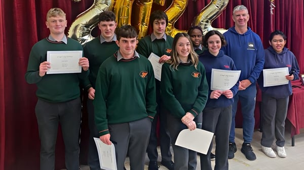 Schüler des St. Fergal’s College stehen in Schuluniformen mit Zertifikaten vor einem gold-lila dekorierten Hintergrund.