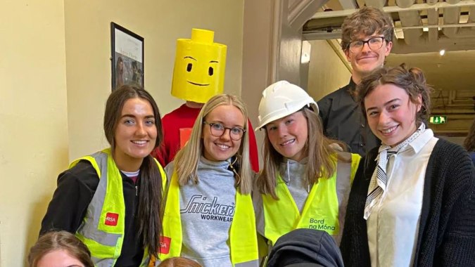 Eine Gruppe von Schülerinnen und Schülern des St. Finian’s College Mullingar trägt Schutzwesten und Helme vor einer großen gelben Smiley-Figur auf einer Baustelle.