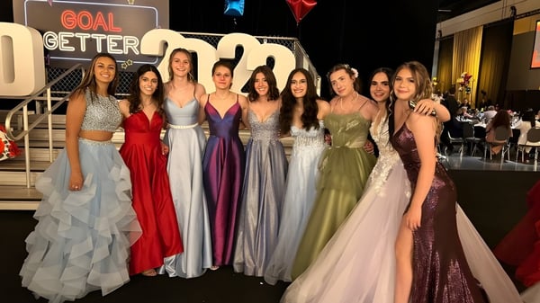 Eine Gruppe junger Frauen in formalen Abendkleidern steht vor einem Hintergrund mit dem Schriftzug GOAL GETTER auf dem Campus der St. Francis High School.