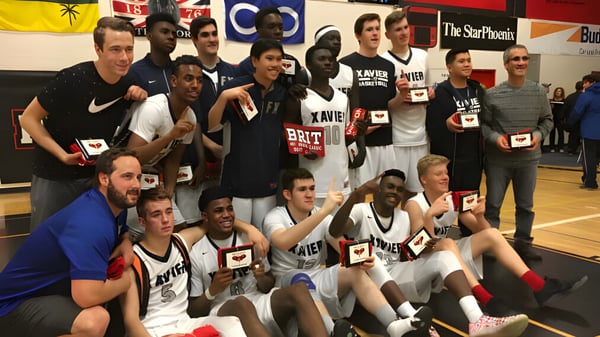 Eine Gruppe von Schülern der St. Francis Xavier High School posiert gemeinsam in einer Sporthalle mit Teamlogos im Hintergrund.