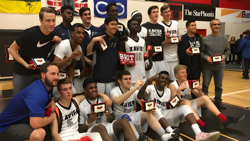 Eine Gruppe von Schülern der St. Francis Xavier High School posiert gemeinsam in einer Sporthalle mit Teamlogos im Hintergrund.