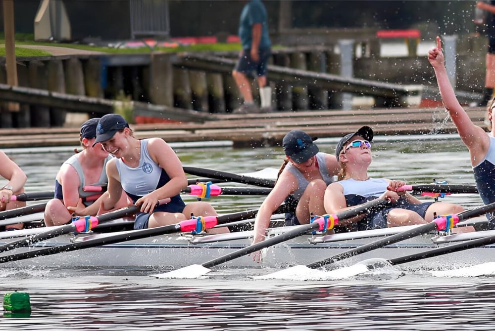 Eine Gruppe von Schülerinnen und Schülern der St. Hilda's Collegiate School rudert auf dem Wasser vor einem Steg und Gebäuden.