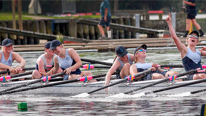Eine Gruppe von Schülerinnen und Schülern der St. Hilda's Collegiate School rudert auf dem Wasser nahe einem Steg und Gebäuden.