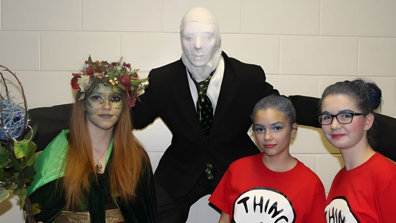 Drei Schülerinnen der St. James' Regional High posieren in farbenfrohen und ausgefallenen Kostümen vor einer gefliesten Wand.