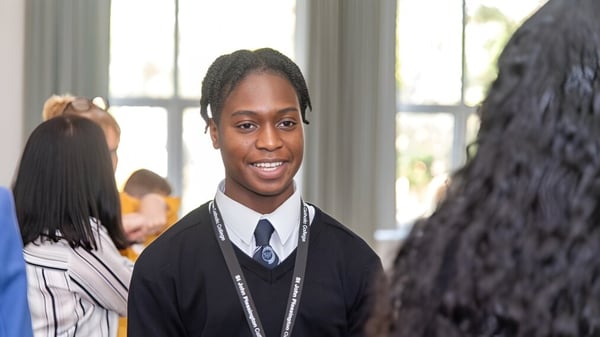 Ein Schüler im Schuluniform steht lächelnd im Klassenraum des St. John Plessington Catholic College mit weiteren Schülern im Hintergrund.