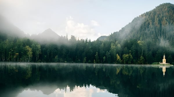 Ein ruhiger See mit einer kleinen Kirche am Ufer, umgeben von grünen Wäldern und Bergen, nahe St. John’s-Kilmarnock School.