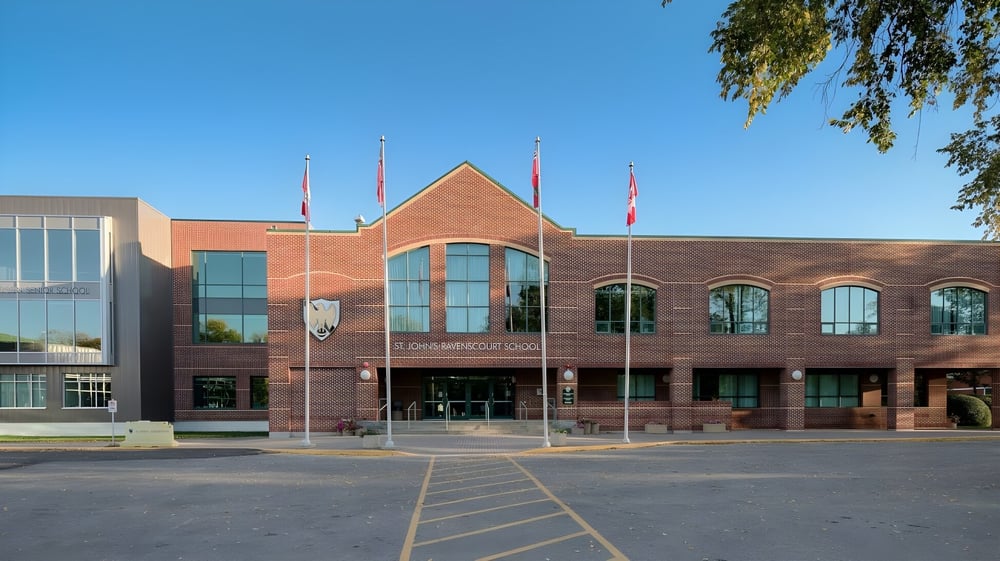 Das große Backsteingebäude mit Rundbogenfenstern auf dem Campus der St. John’s-Ravenscourt School ist von gepflastertem Bereich und Bäumen umgeben.