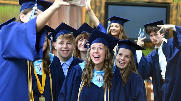 Eine Gruppe von Absolventinnen und Absolventen der St. John’s-Ravenscourt School in blauen Abschlussroben stehen zusammen im Freien vor einem Holzzaun.