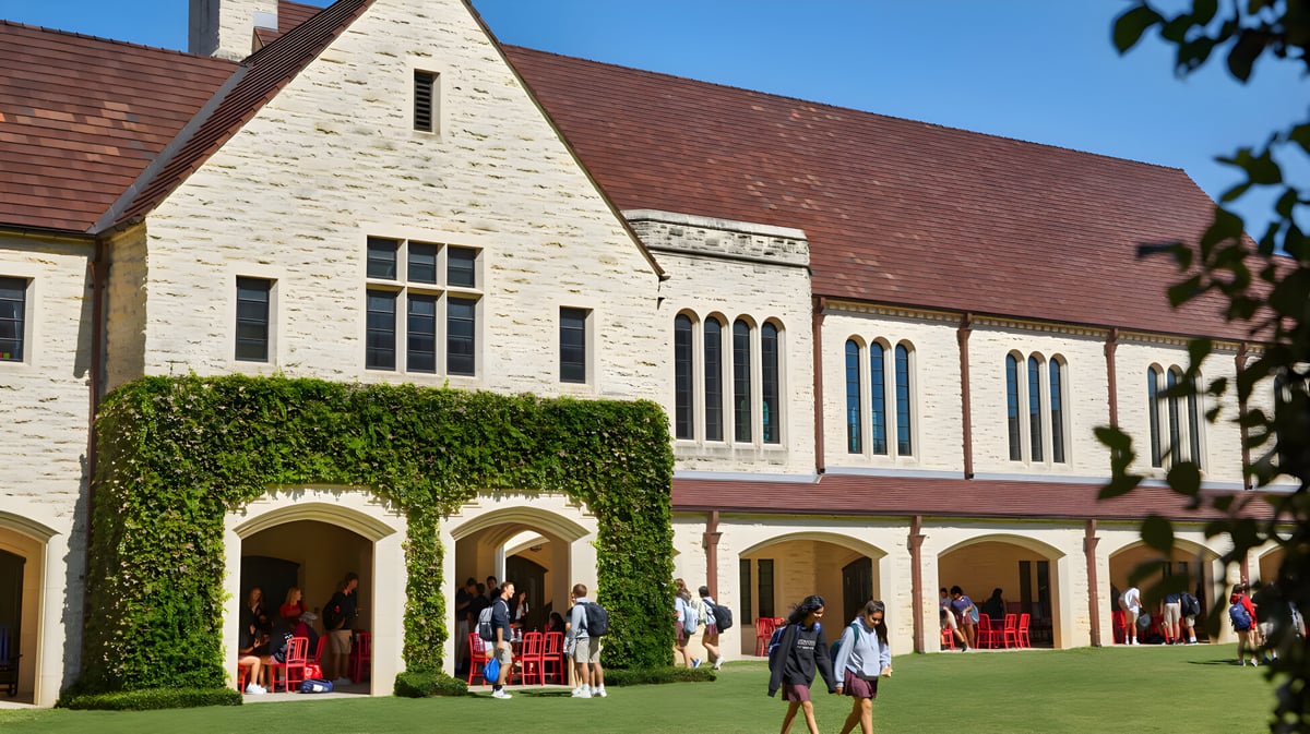 Das große Steingebäude der St. John's School ist von üppigem Grün umgeben und im Vordergrund sitzen Menschen auf Bänken.