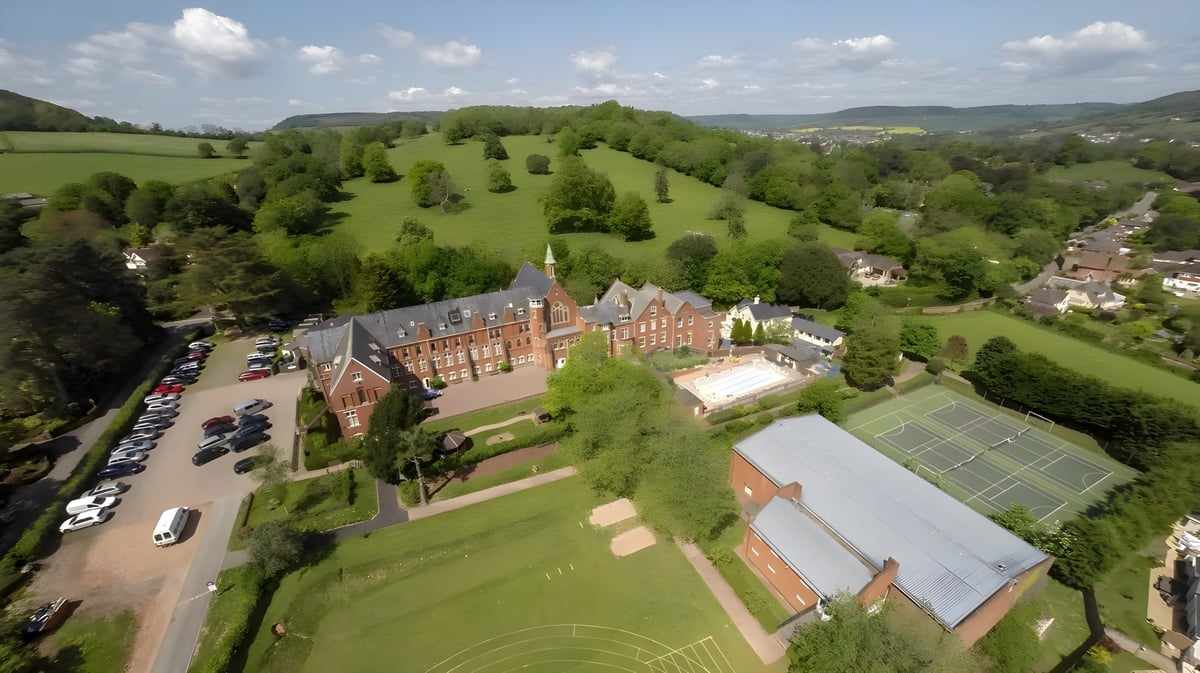 Das historische Gebäude der St. John’s School Sidmouth liegt eingebettet in grüne Hügel und Waldlandschaften mit einem Tennisplatz im Vordergrund.