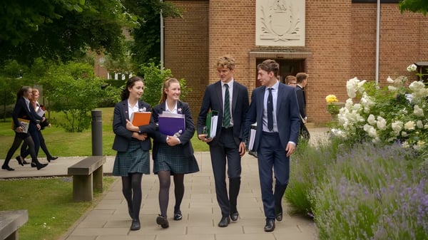 Eine Gruppe von Schülern geht gemeinsam auf einem Weg mit grüner Umgebung vor einem Backsteingebäude auf dem Campus der St. John’s School in Leatherhead.