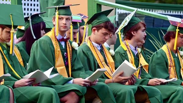 Eine Gruppe von Absolventinnen und Absolventen der St. Johnsbury Academy in grünen Talaren steht mit Diplomen vor einem bunten Hintergrund.
