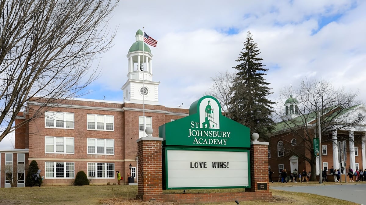 Das Backsteingebäude mit Uhrturm und amerikanischer Flagge auf dem Campus der St. Johnsbury Academy ist von Bäumen und einer Wiese umgeben.