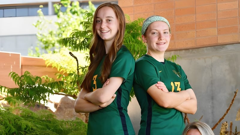 Zwei Schülerinnen der St. Joseph Catholic High School stehen in grünen Trikots vor einem Backsteingebäude mit Grünpflanzen.