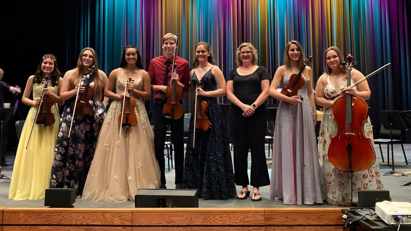 Schüler der St. Joseph High School stehen in formeller Kleidung mit Musikinstrumenten auf einer Bühne vor farbigem Hintergrund.