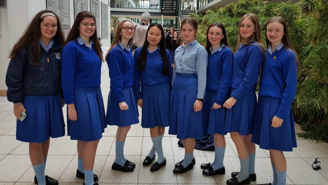 Eine Gruppe von Schülerinnen der St. Joseph of Cluny School steht in einheitlichen blauen Uniformen in einem gefliesten Innenraum mit Glaswand.