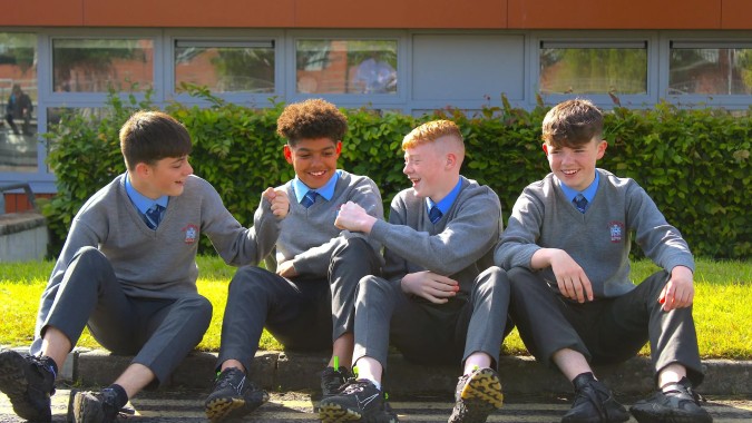 Vier Schülerinnen und Schüler der St. Joseph's Secondary School sitzen auf einer Bank vor dem Schulgebäude mit großen Fenstern und Grünflächen.