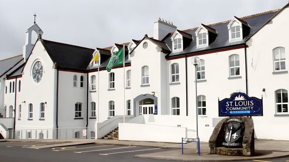 Das mehrstöckige weiße Gebäude mit einem kirchturmähnlichen Turm und Fahnen auf dem Gelände der St. Louis Community School mit einem Steindenkmal im Vordergrund.