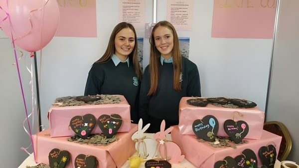 Zwei Schülerinnen der St. Louis Secondary School stehen vor einem rosa Hintergrund mit zwei großen Kuchen mit Herzdekor in der Front.