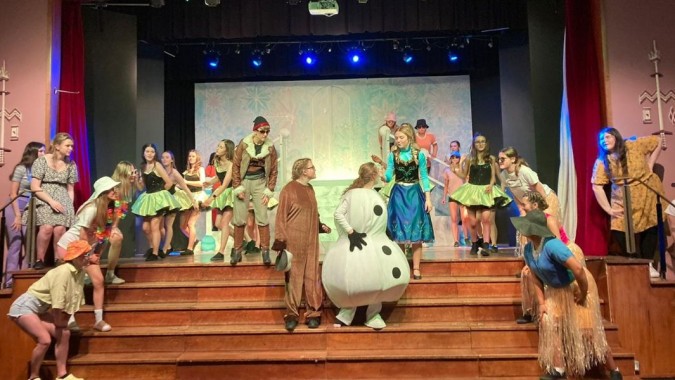 Eine Gruppe von Schülern der St. Louis Secondary School steht in bunten Kostümen mit einer großen Schneemannfigur auf der Bühne vor einem winterlichen Bühnenbild.