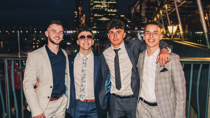 Vier Schüler des St. Macartan's College stehen in formeller Kleidung vor der nächtlichen Stadtkulisse mit hohen Gebäuden.