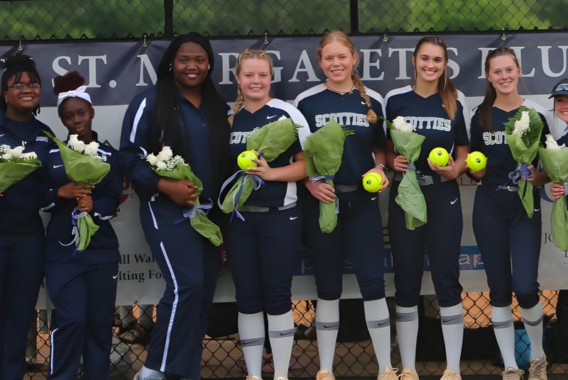 Eine Gruppe von Softballspielerinnen in Uniform steht vor einem Zaun mit einem Banner der St. Margaret's School.