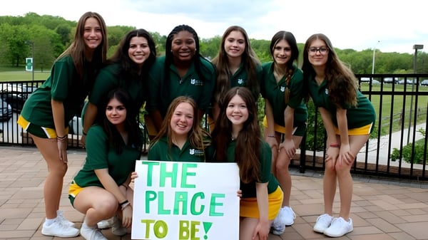 Eine Gruppe Schülerinnen der St. Mark High School steht mit einem Schild 'The Place to Be' vor einem Park mit Bäumen und Zaun.