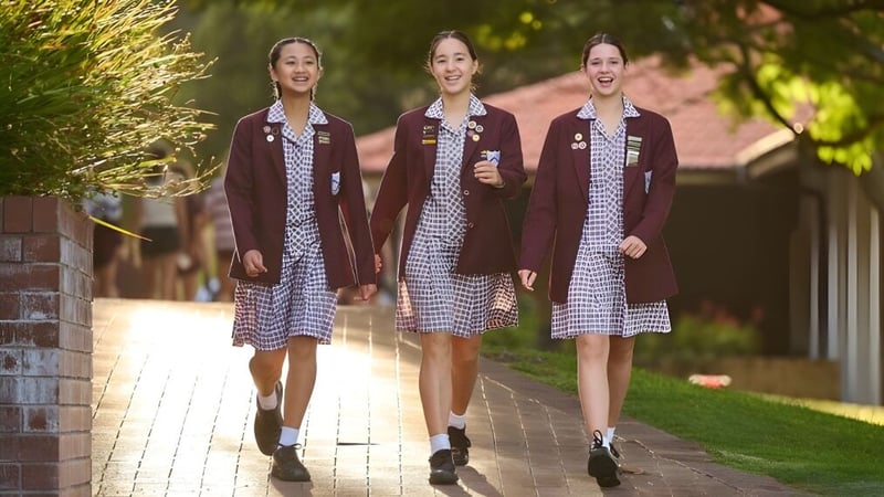 Drei Schülerinnen der St. Mary's Anglican Girls' School gehen auf einem Weg zwischen Grünflächen und Backsteingebäuden.