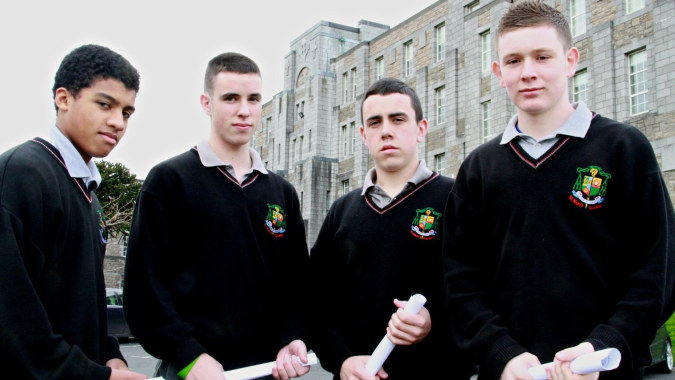 Vier Schülerinnen und Schüler in schwarzer Robe mit Wappen stehen vor einem Steingebäude mit Rundbogenfenstern auf dem Campus von St. Mary’s College Ballysadare.