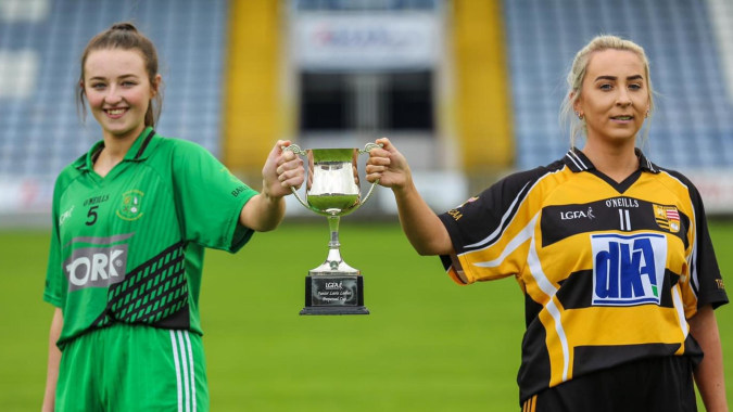 Zwei Fußballspielerinnen der St. Mary's Holy Faith Killester halten gemeinsam einen Pokal auf dem Fußballfeld.